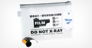 A mesh zipper pouch labeled "FILM DO NOT X-RAY" in English and other languages, with a graphic of a film roll and airport security icons, a travel tag, and a film canister placed in front of the pouch.