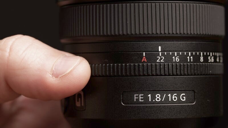 Close-up of a hand adjusting the focus ring on a camera lens labeled "FE 1.8/16 G." The aperture scale is visible, showing settings from f/1.8 to f/22, with the focus set to A. The lens surface is matte black with textured grips.