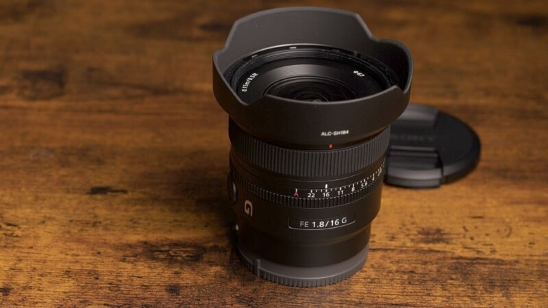 A black camera lens with a lens hood attached sits on a wooden surface. The lens cap is placed nearby. The focus ring and aperture markings are visible, indicating specifications.