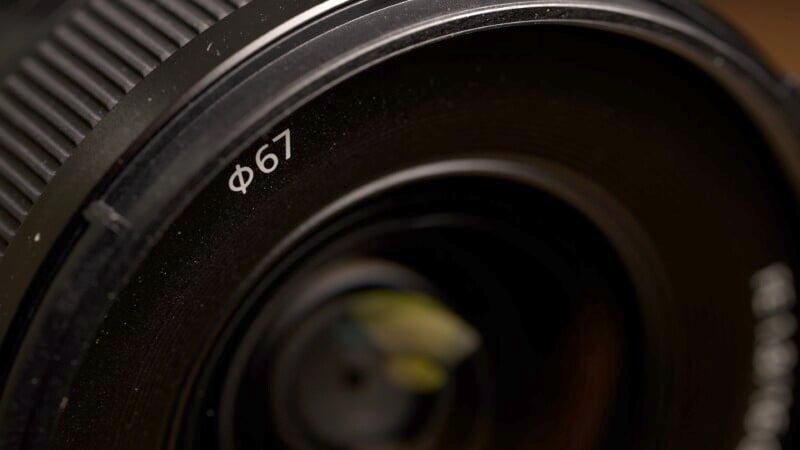 Close-up of a camera lens with a black exterior and ridged focus ring. The marking "Φ67" is visible on the lens, indicating a 67mm filter size. The lens reflects light, revealing part of the internal glass elements.