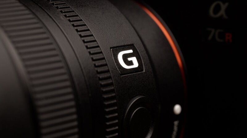 Close-up of a camera lens with a prominent "G" logo, featuring a textured focus ring and part of the camera body visible, highlighted by soft lighting.