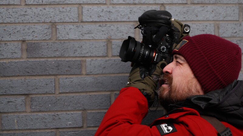 A person in a red jacket and maroon beanie is holding a camera up to their eye, aiming at a gray brick wall. The person is wearing green gloves and appears focused on taking a photo.