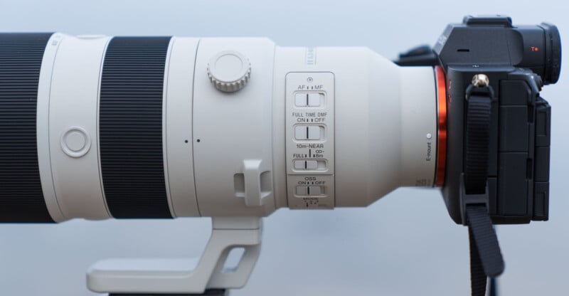 Close-up of a white telephoto camera lens attached to a black camera. The lens features multiple switches for autofocus and stabilization settings. It is mounted on a tripod collar. The setup is set against a plain, light background.