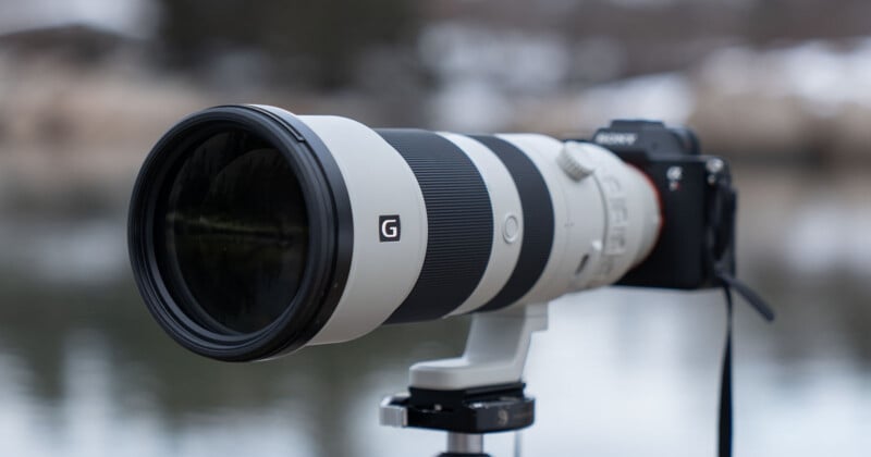 A professional camera with a large telephoto lens mounted on a tripod. The lens is prominently black and white with the letter "G" on it, set against a blurred outdoor background.