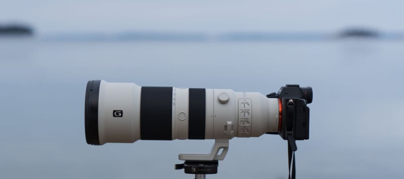 A large telephoto camera lens mounted on a camera sits on a tripod against a blurred, serene landscape, featuring a calm body of water and distant islands or landforms in the background.
