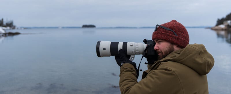 A person in a red knit hat and green jacket uses a large camera lens to photograph a tranquil body of water. The backdrop features a cloudy sky, calm water, and distant trees.