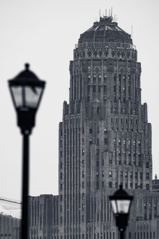 Black and white photo of a tall, intricate Art Deco skyscraper. In the foreground, two blurred street lamps frame the building. The sky is overcast, enhancing the dramatic, vintage feel of the architectural scene.