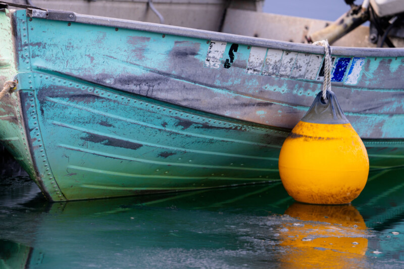 A close-up of a weathered green boat with peeling paint, floating on a calm body of water. A bright yellow buoy hangs from the side, partially submerged, reflecting in the water's surface.