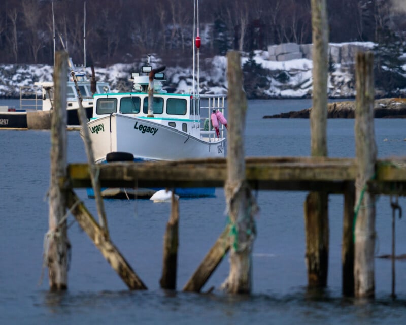 A white fishing boat named "Legacy" is moored on calm water near a rustic wooden dock. The dock is in the foreground, and a snowy shoreline with trees is visible in the background.
