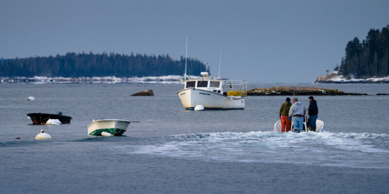 Three people in a small motorboat travel toward a larger, anchored fishing boat on a calm body of water. Several buoys and small boats are visible, with a forested shoreline in the background under a cloudy sky.