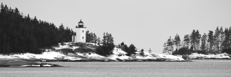 Black and white image of a coastal scene featuring a lighthouse on a snowy cliff surrounded by pine trees. The foreground shows calm water, while the lighthouse stands tall, guiding near the densely wooded shoreline.