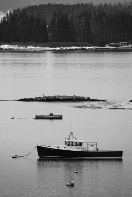 A black and white image of a small boat anchored on a calm lake with buoys nearby. In the background, there's a small island and a forested shoreline with trees. The water reflects the overcast sky.