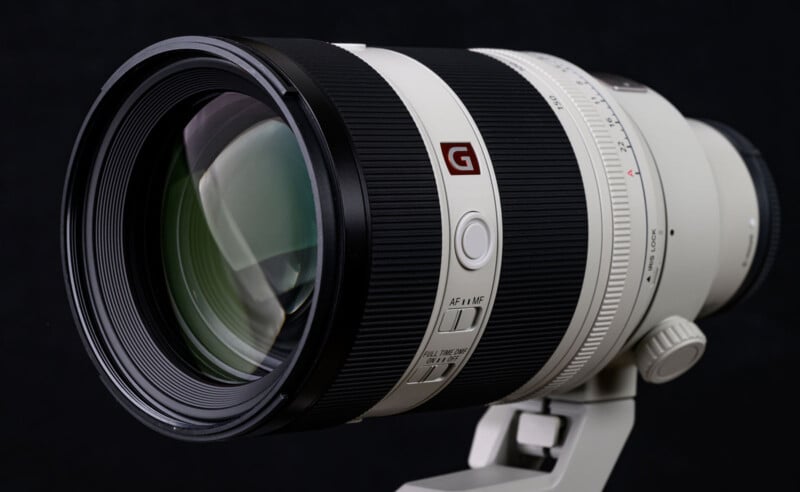 A close-up of a large white telephoto camera lens with a prominent glass element, black grip rings, control switches, and a red "G" logo, mounted on a tripod collar against a black background.