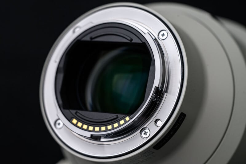 Close-up of the rear element and electronic contacts of a camera lens, with a metallic mount and glass optics visible, set against a dark background.