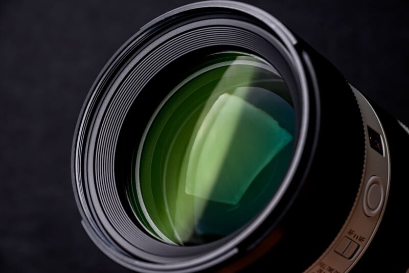 Close-up view of a camera lens with green reflections on the glass elements, set against a dark, blurred background. The detailed rings and buttons around the lens are also visible.