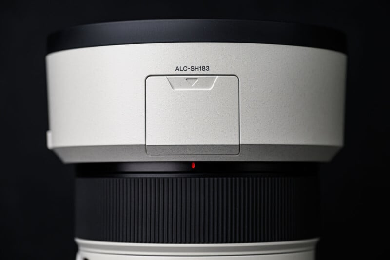 Close-up of a camera lens with a white and black finish, labeled "ALC-SH183" near the top. The lens features a textured focus ring and a small red alignment dot below a rectangular panel.