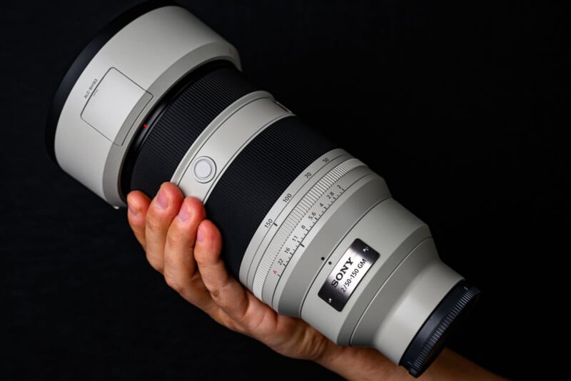 A hand holding a large Sony camera lens with white and black detailing against a black background. The lens shows measurement markings and a Sony logo with "FE 400mm F2.8 GM OSS" label.