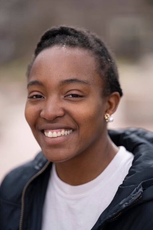 A person with short hair smiles warmly at the camera, wearing a black jacket, white shirt, and small stud earrings. The background is softly blurred, keeping the focus on their face.