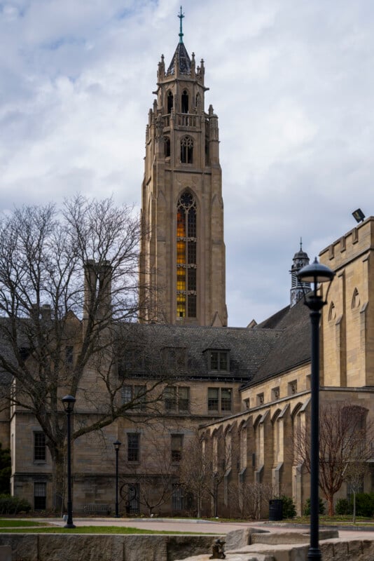 A tall, ornate Gothic-style bell tower rises above a stone university building with leafless trees and lamp posts in the foreground under a cloudy sky.