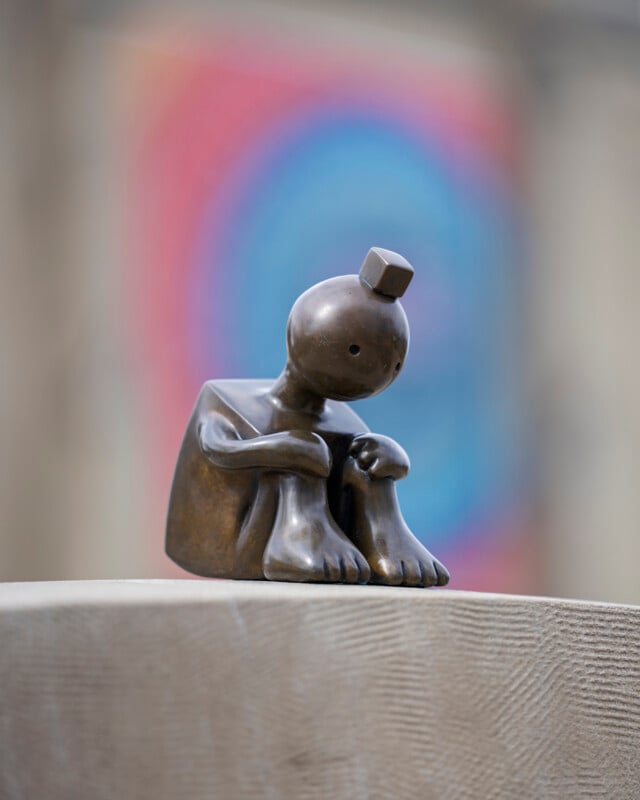 A bronze sculpture of a stylized, seated figure with a square body, large feet, and a small round head topped with a cube, sitting with knees drawn up on a textured pedestal; colorful blurred background.