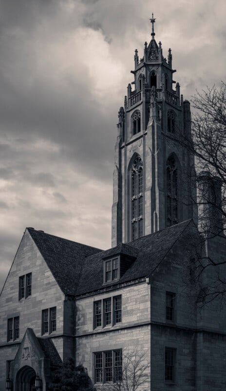 A tall, ornate stone church tower rises above a gothic-style building under a cloudy sky, with bare tree branches visible to the right. The scene is in black and white, evoking a dramatic, historic atmosphere.