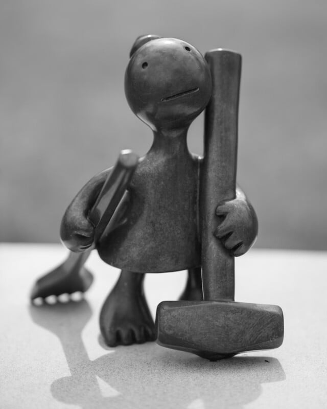 A small, smooth, abstract sculpture of a figure holding a large hammer and another tool, standing on a flat surface with a blurred background.