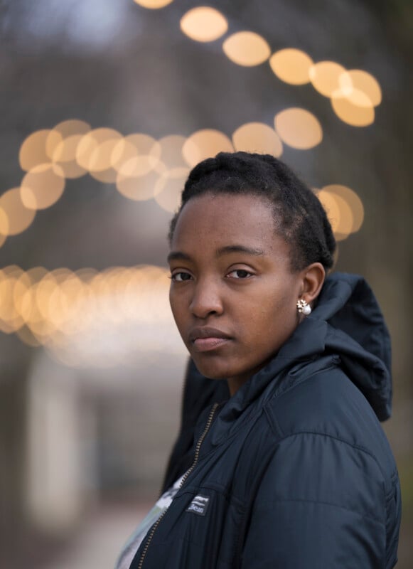 A person with short hair and a serious expression stands outdoors, wearing a dark jacket. Out-of-focus string lights create a bokeh effect in the background.