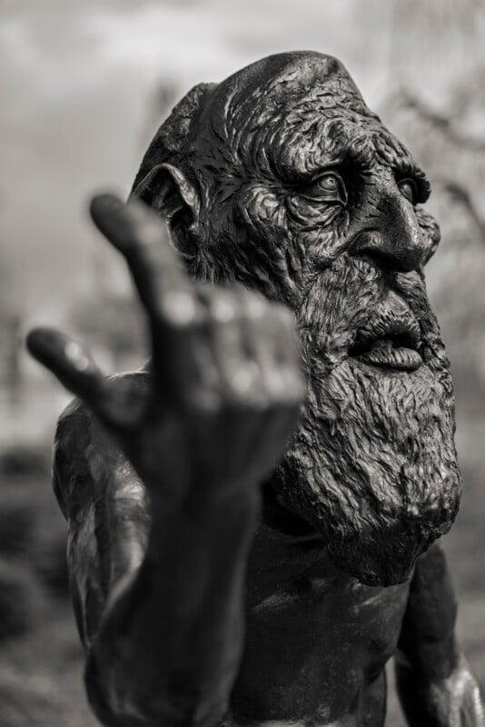 A close-up of a weathered bronze statue of an elderly bearded man, with a deeply lined face and hand outstretched toward the viewer. The background is blurred, emphasizing the statue’s detailed expression.