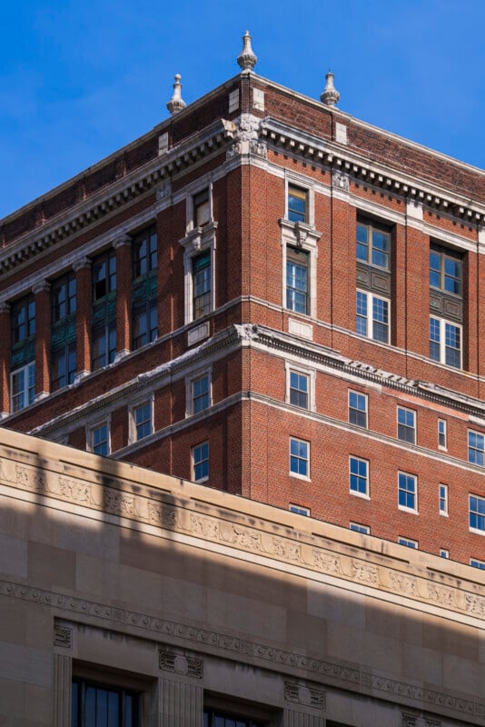 A multi-story red brick building with decorative stone trim rises behind a beige stone structure, both set against a clear blue sky. The brick building features tall windows and ornate architectural details.