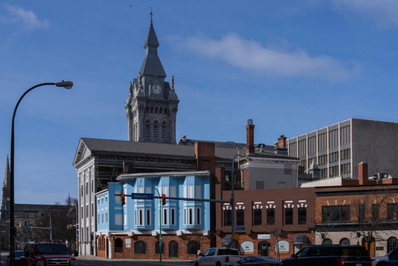 A downtown street scene shows historic buildings, including a tall clock tower, under a blue sky. Traffic lights hang above the intersection with cars stopped in front of colorful storefronts and brick façades.