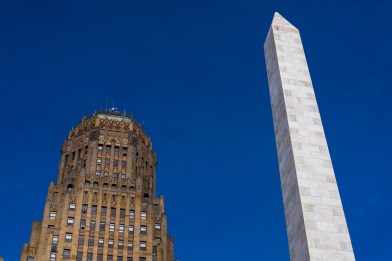 A tall, ornate brown building with Art Deco details stands next to a white marble obelisk monument against a clear blue sky.