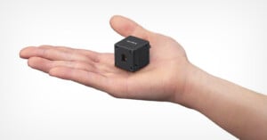 A small black cube-shaped camera rests on the open palm of a person’s hand, highlighting the device’s compact size. The background is plain and light colored.