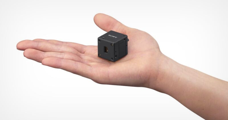 A small black cube-shaped camera rests on the open palm of a person’s hand, highlighting the device’s compact size. The background is plain and light colored.