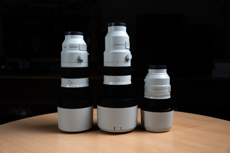 Three camera lenses of varying sizes stand upright on a wooden surface against a dark background. Each lens is white with black and silver accents, featuring various dials and switches for adjustments.