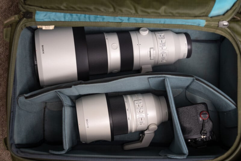 Two large Sony camera lenses are placed inside a padded camera backpack. The lenses are secured in individual compartments. A small black camera body is partially visible in the bottom right section of the bag.