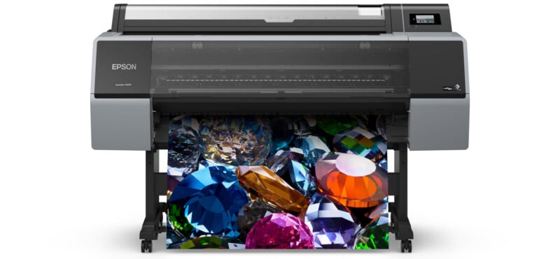 A large Epson printer prints a vibrant, high-resolution image of various colorful gemstones, including blue, orange, pink, and green gems, on a sheet of paper.
