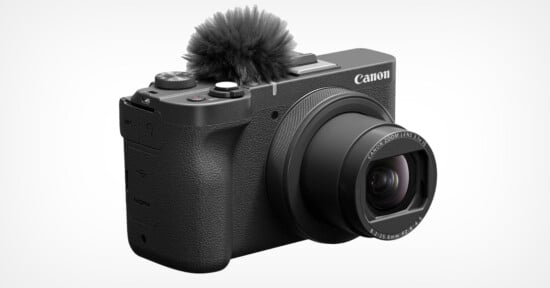 A Canon digital camera with a large lens and a built-in windscreen over the microphone, viewed at an angle. The camera body is textured, and various control buttons and dials are visible on the top and side.