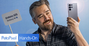 A person with a beard holds up a smartphone, smiling and facing the device. They stand against a blue backdrop. A sign next to them reads "Galaxy S25 Galaxy AI" and "PetaPixel Hands-On.