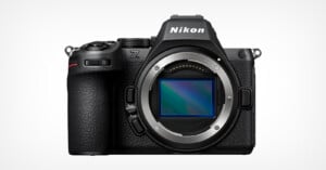 A Nikon Z series mirrorless camera body is displayed against a white background. The camera has a visible lens mount without a lens attached, showcasing its sensor. It features a black textured grip and various buttons and dials.