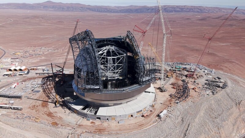 Aerial view of a large telescope under construction in a barren desert, surrounded by cranes and construction equipment, with distant mountains in the background.