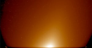 A reddish-brown sky filled with stars above a curved horizon, with a bright, glowing light at the center of the horizon, creating a soft gradient effect.