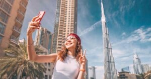A smiling woman wearing sunglasses and a headband takes a selfie and makes a peace sign, with tall modern buildings and the Burj Khalifa visible in the background under a clear blue sky.