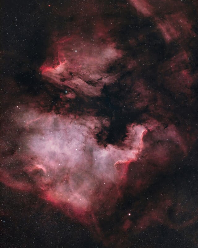 A vivid photograph of the North America Nebula in space, showing clouds of glowing pink and red gas, dark dust lanes, and scattered stars against a black background.