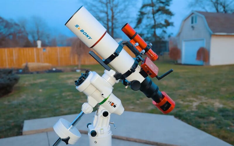 A white and red telescope mounted on a tripod stands outdoors on a concrete patio, with a grassy yard, wooden fence, trees, and a small shed visible in the background.