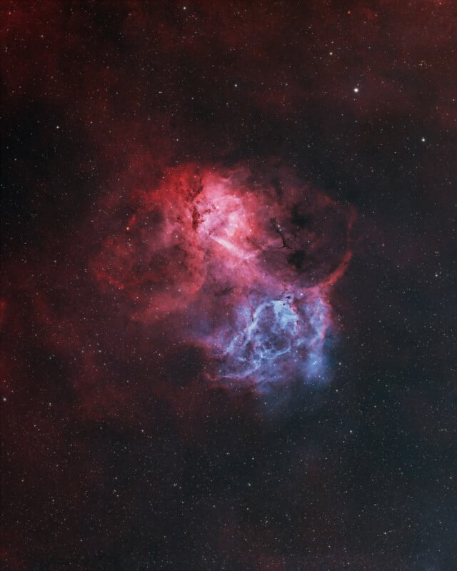 A vibrant nebula in deep space, glowing with red and blue hues surrounded by scattered stars on a dark background. The cloud-like structure has intricate, wispy details and contrasts against the blackness of space.