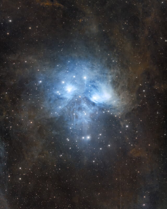 A bright blue nebula with wispy clouds of gas and dust glows against a dark background, surrounded by numerous stars scattered across space.