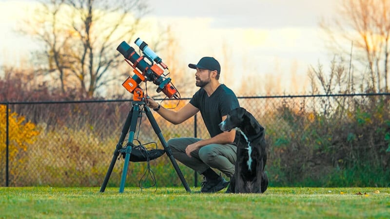A man kneels on grass beside a black and white dog, adjusting a large telescope on a tripod outdoors, with a fence and trees in the background.