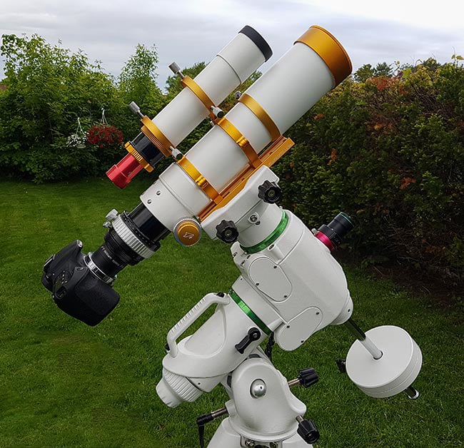 A white and gold refractor telescope with a camera attached, mounted on a sturdy equatorial mount, set up outdoors on a grassy lawn with bushes and trees in the background.