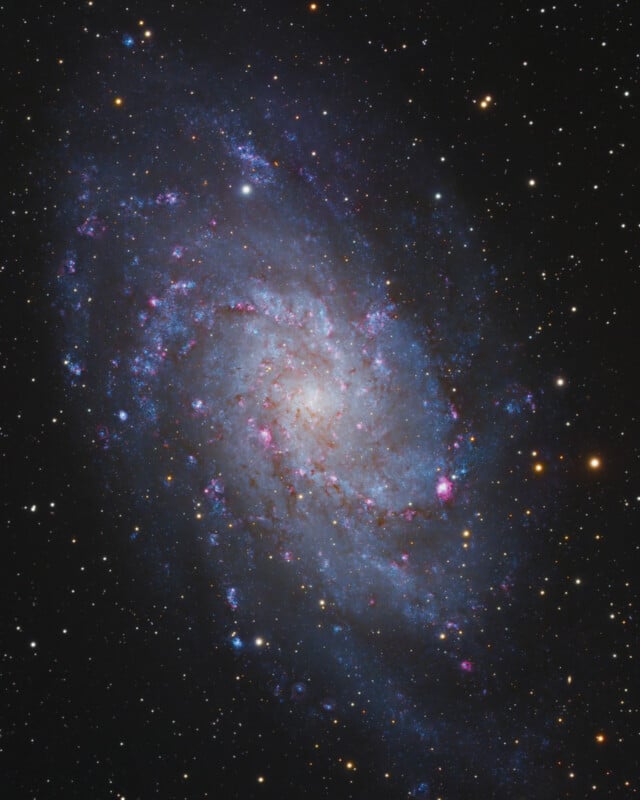 A detailed image of a spiral galaxy, showing bright, swirling blue arms with pink star-forming regions and a glowing core, surrounded by numerous distant stars against a dark background.