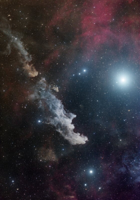 A deep space image shows the Witch Head Nebula, a blue-tinted, wispy cloud of gas and dust, surrounded by countless stars and a bright, glowing star in the upper right corner. Red and dark cosmic clouds fill the background.